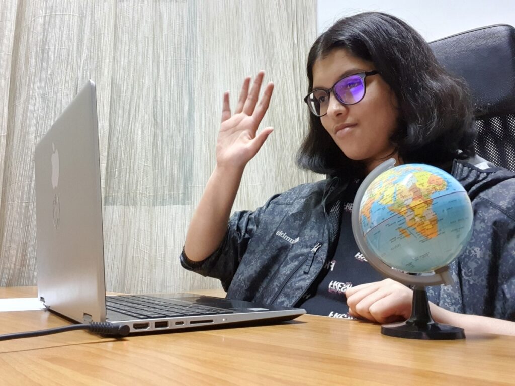 A Junior Academy participant sits at a desk in front of an open laptop, raising her hand and participating in a virtual video call.