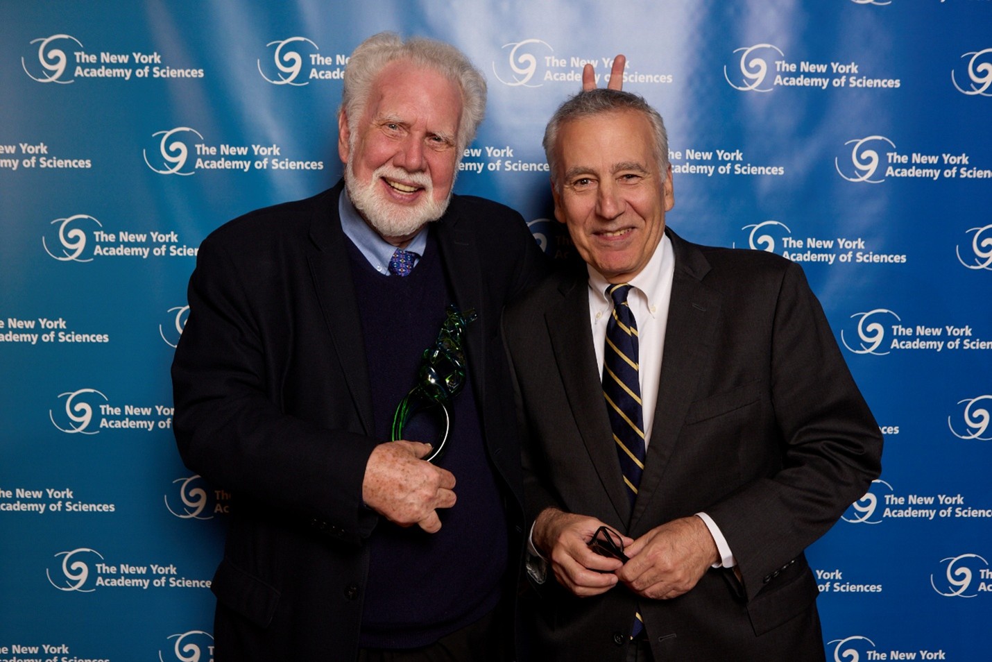 Two formally dressed men pose together. The one man gives his friend bunny ears while goofing around.